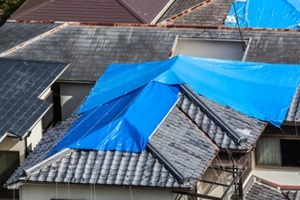 view of houses hit by natural disaster, with damaged tiled roof covered with blue tarp